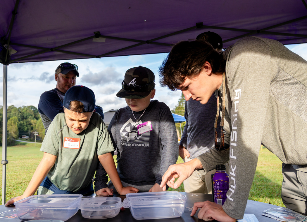 Several people gathered under a purple canopy table outdoors, looking at clear plastic containers on a table. One person wears a gray long-sleeve shirt with vertical white text reading 'NUKIFISHING'.
