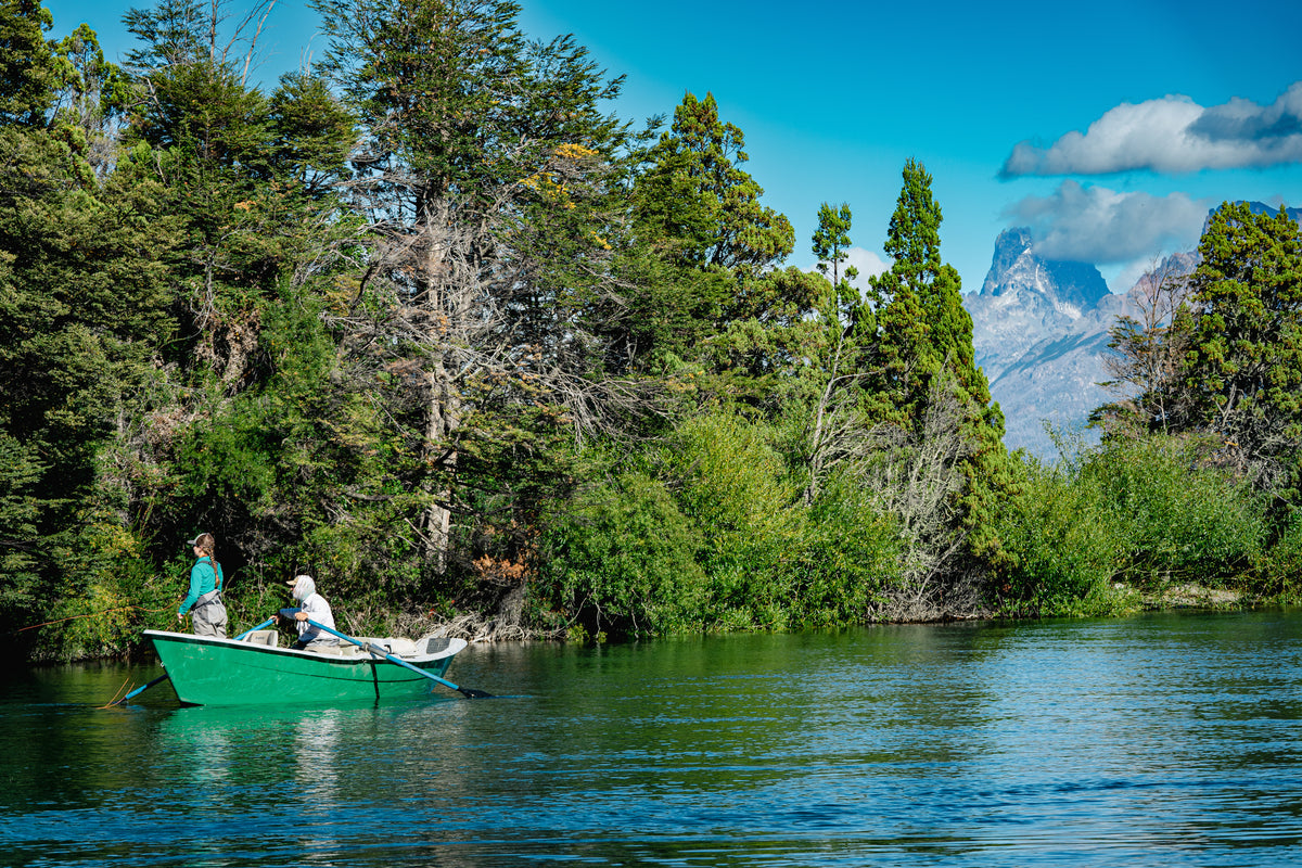 A scenic outdoor photograph showing a small green rowboat on calm blue water, with two people in the boat and dense green trees lining the shore. A mountain peak is visible in the background under a bright blue sky with a few clouds.