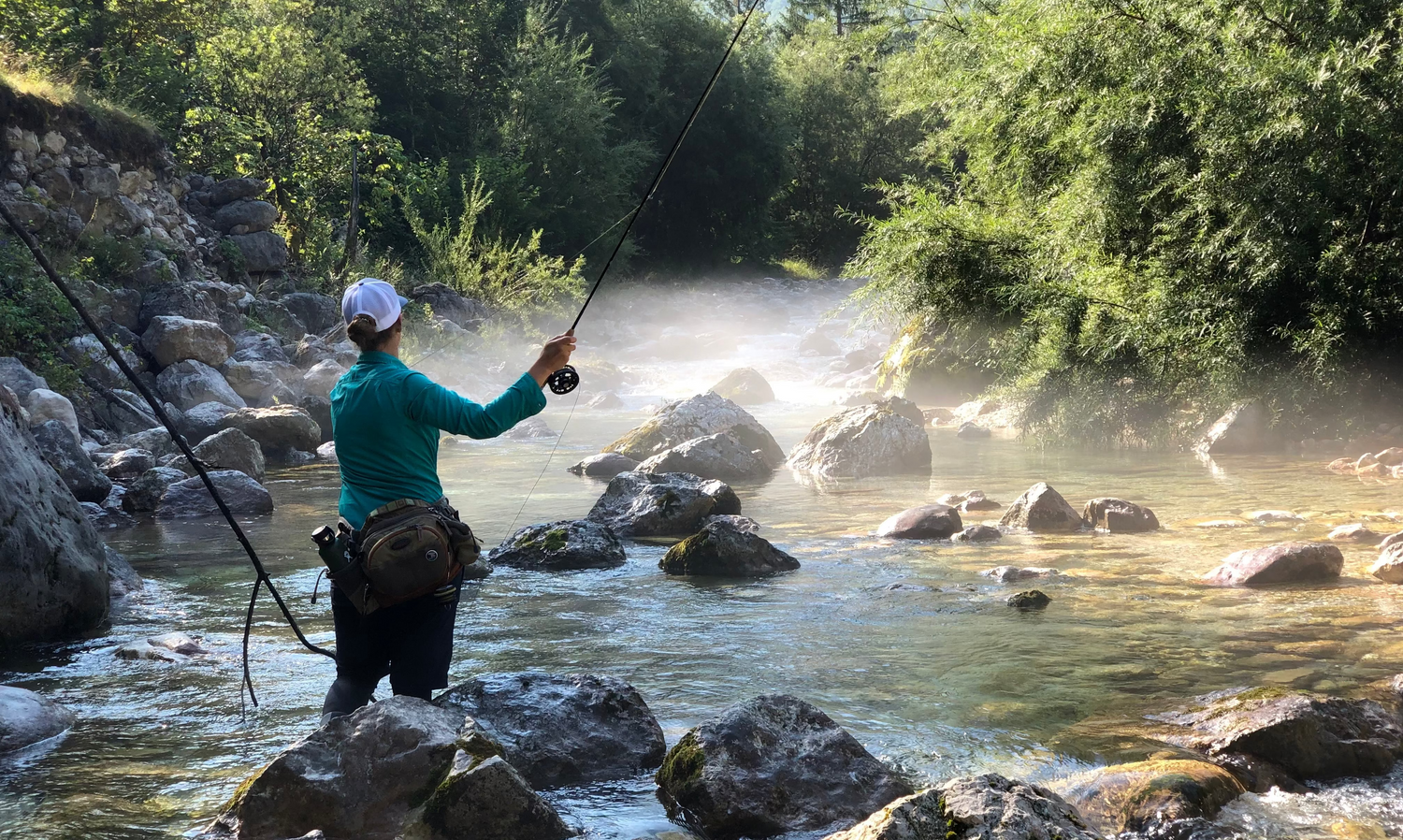 A person fly fishing in a rocky river with mist in the background, surrounded by lush green forest. The person is wearing a teal long-sleeve shirt, black pants, and a white cap while holding a fishing rod.